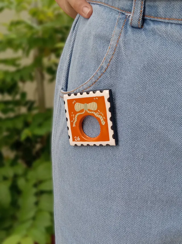 Studio Gayatri handmade boho orange postage stamp clay sunglasses holder brooch with a hand-painted bow detail.