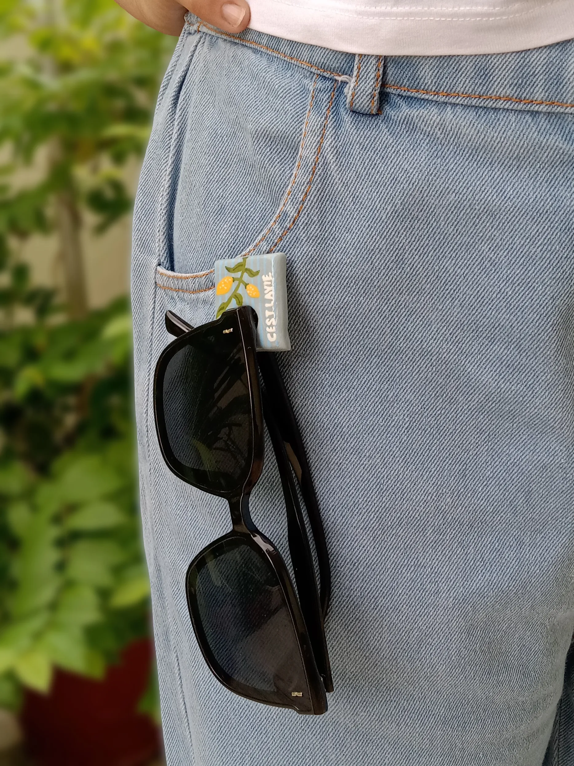 A pair of sunglasses hanging securely from the loop of a hand-painted lemon branch clay brooch pinned to denim jeans