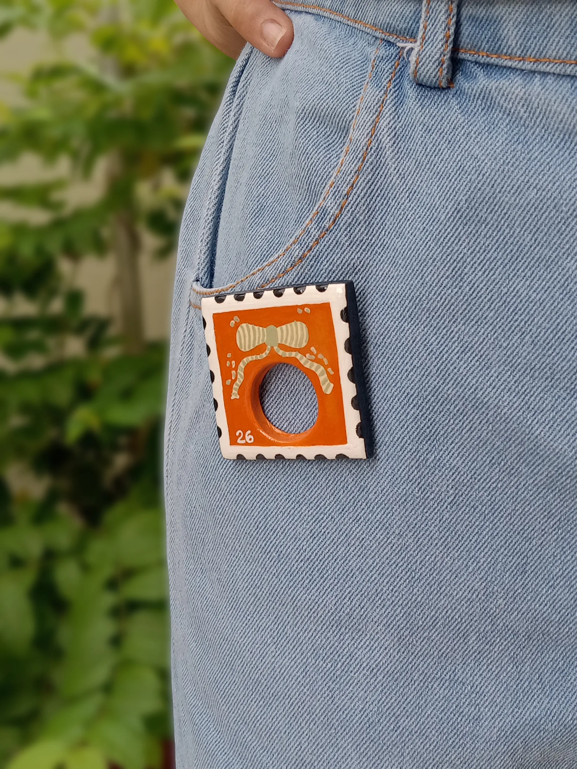 Studio Gayatri handmade boho orange postage stamp clay sunglasses holder brooch with a hand-painted bow detail.