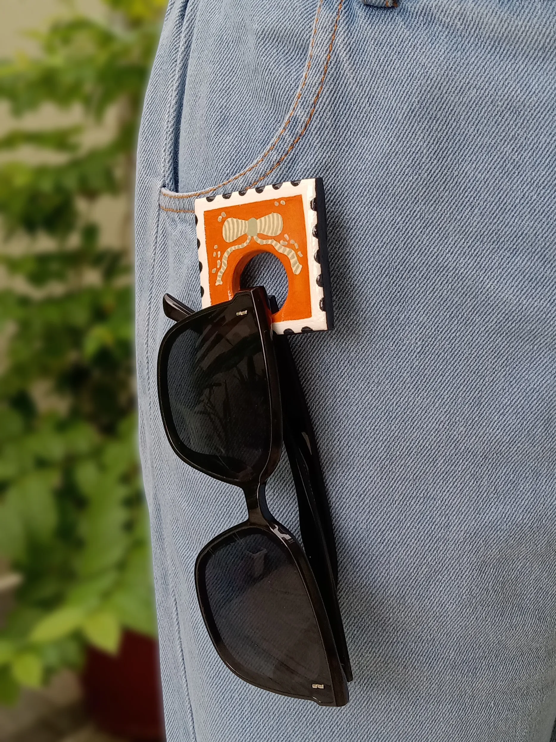 Close up of sun glasses hanging securely from the loop of an orange clay postage stamp brooch pinned to a denim pocket.